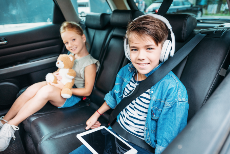 Kids in the car with a boy wearing headphones and using a tablet.