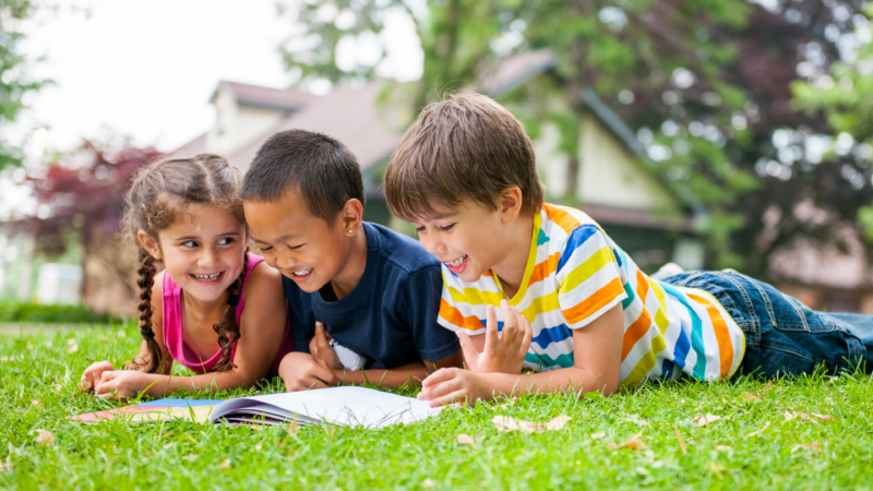 Three children reading a book together outdoors, representing fun summer reading activities for kids with Epic Books for Kids.