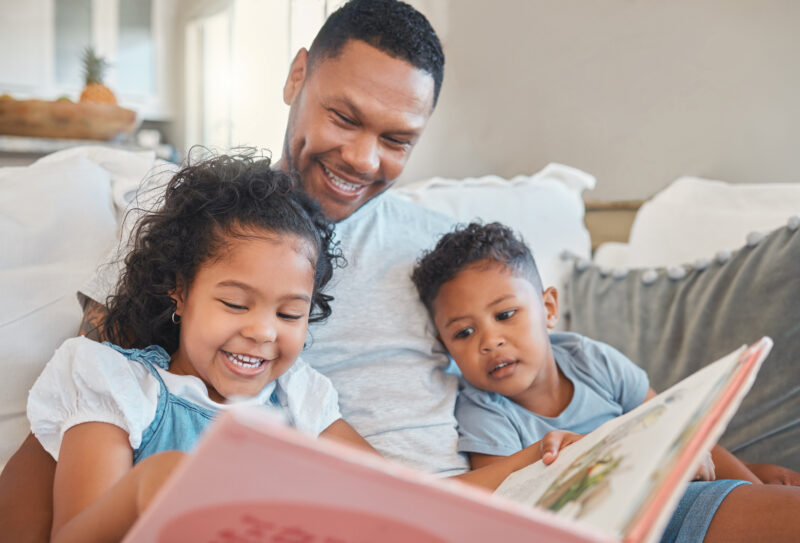 Everyone gets a turn to read. Shot of a young man reading to his kids on the sofa at home.